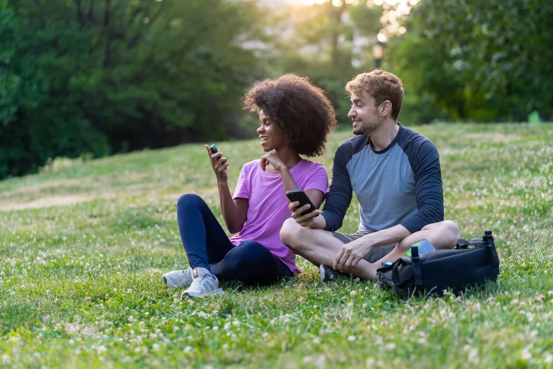 Femme assise sur l’herbe, tenant un vaporisateur NICORETTE VapoÉclair®, à côté d’un homme qui a son téléphone à la main